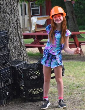 Young white girl wearing a hard hat getting ready to toss a milk crate to another camper at Camp Kupugani in Illinois.
